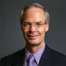 Headshot of Ethan Lazarus, MD, FOMA, sitting in front of a gray background with a black suit and purple shirt