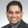 Headshot of Peminda K Cabandugama MD, DABOM, in front of a light gray background, wearing a black suit and blue and black striped shirt.