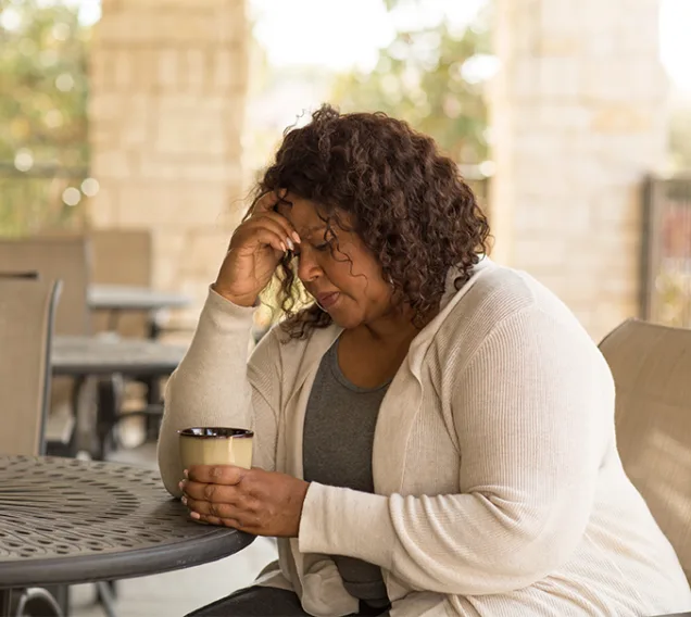 woman sitting on a patio at a table with a cup of coffee, with her head in her hands