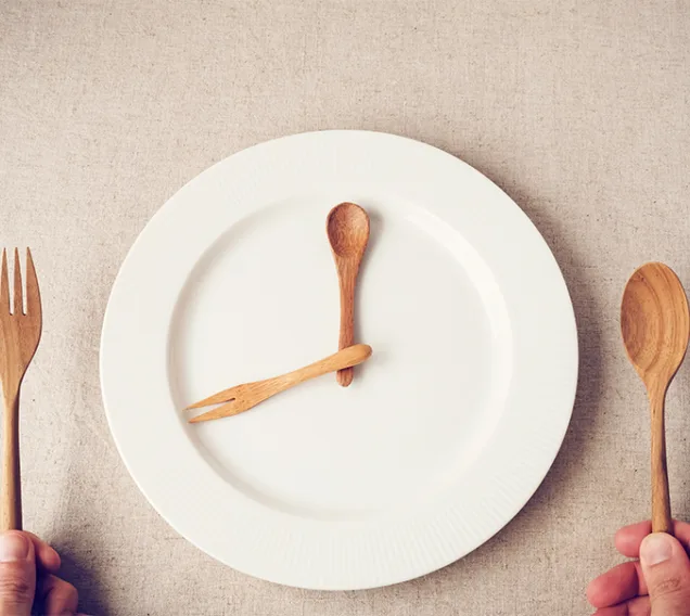 Image of a plate with a person holding a fork and spoon on either side of the plate. Another fork and spoon sit on the plate, to look like clock hands, making the plate look like a clock
