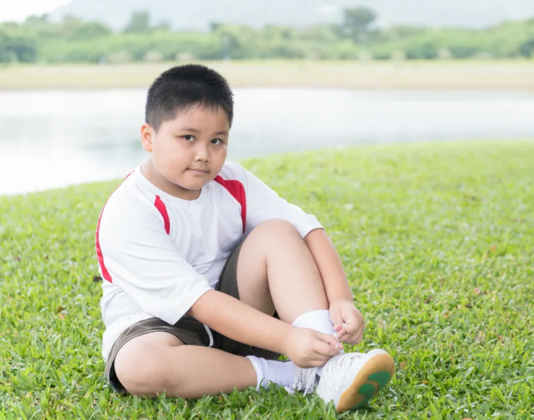 Image of a young boy sitting on the grass tying his shoe and smiling at the camera
