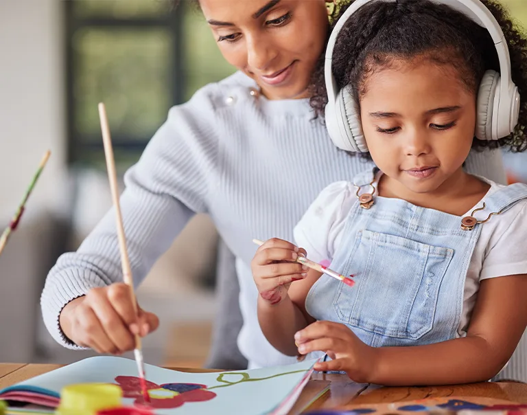 Image of a woman with a child in her lap wearing headphones. Both are painting at a table in front of them, smiling at the artwork.