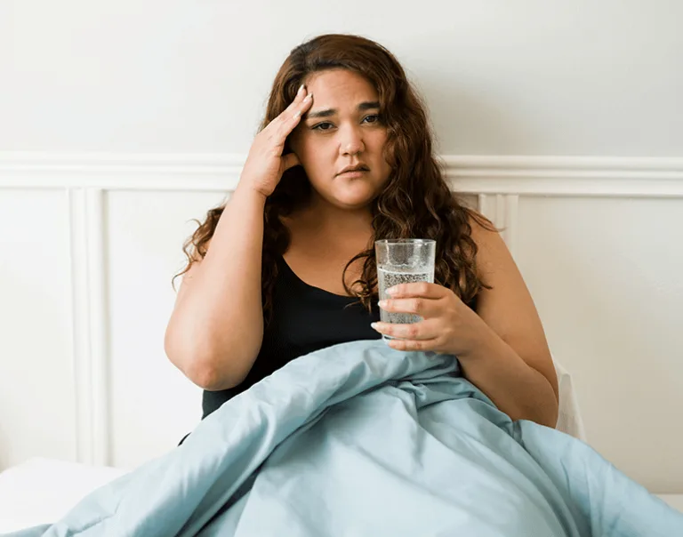image of a young woman sitting in bed with a glass of water, holding her head in pain