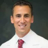 Headshot of Anthony Auriemma, MD, JD, FOMA, posing in front of a gray background, wearing a white coat with a white undershirt and red tie.