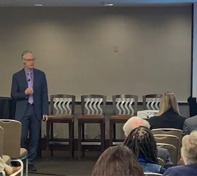 Image of Dr. Ethan Lazarus speaking in front of an audience in a conference room, with a presentation on the screen next to him