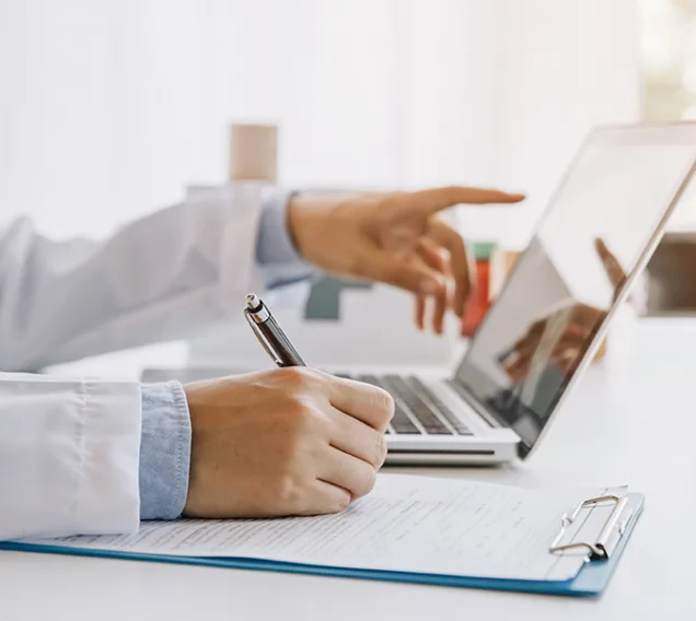 Image of a doctors hands writing on a notepad and pointing at a computer