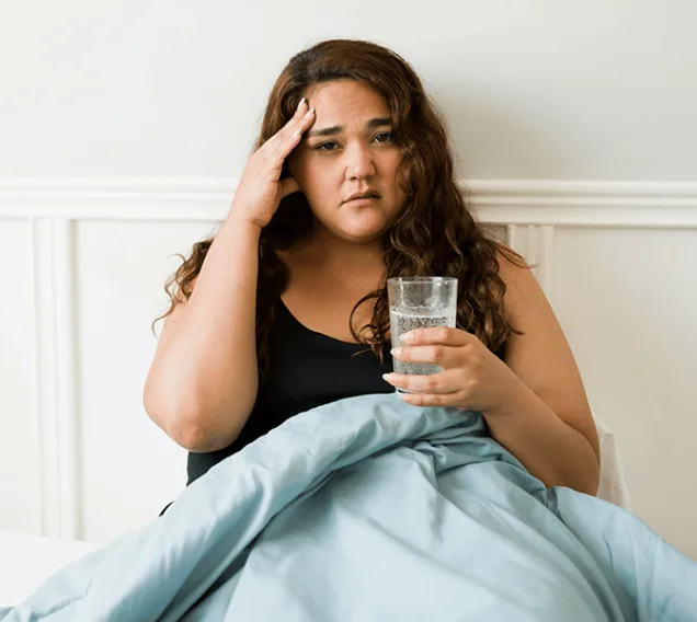 image of a young woman sitting in bed with a glass of water, holding her head in pain