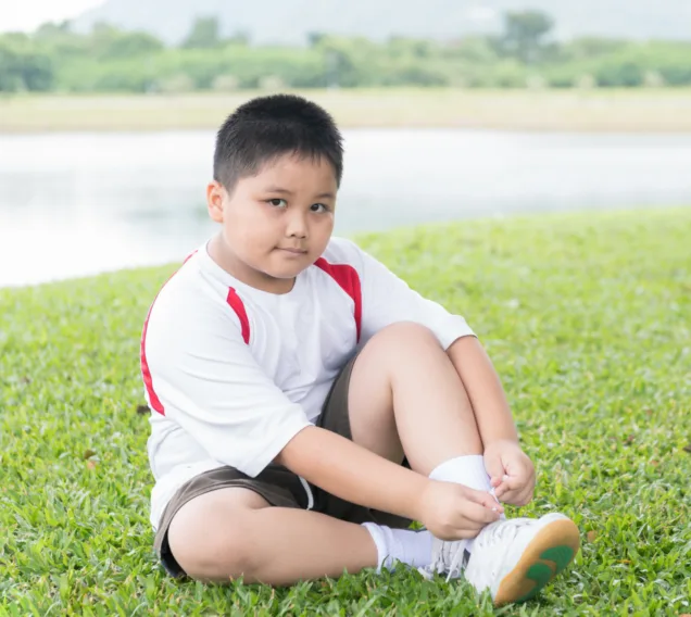 Image of a young boy sitting on the grass tying his shoe and smiling at the camera