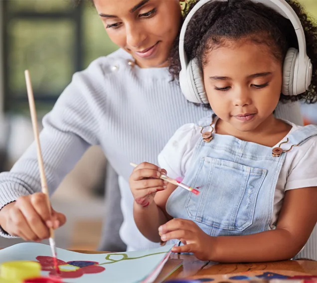 Image of a woman with a child in her lap wearing headphones. Both are painting at a table in front of them, smiling at the artwork.