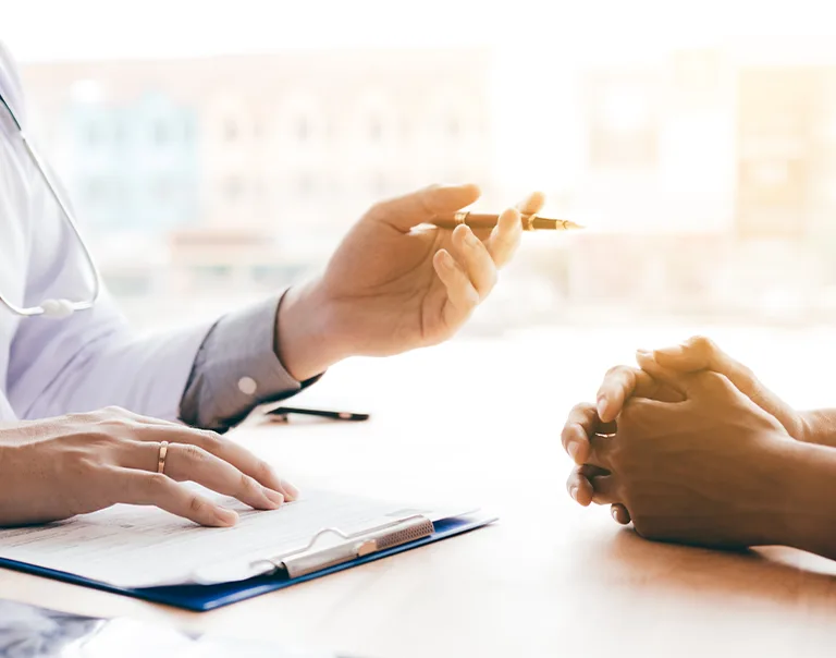 Image of a doctor speaking with a patient over a table with a patient with only their hands in the frame. The doctor is wearing a white coat with a stethoscope and a clipboard on the table.
