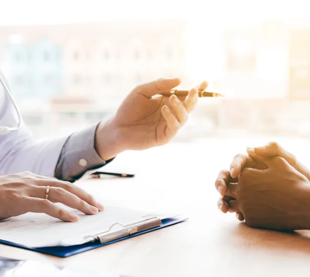 Image of a doctor speaking with a patient over a table with a patient with only their hands in the frame. The doctor is wearing a white coat with a stethoscope and a clipboard on the table.
