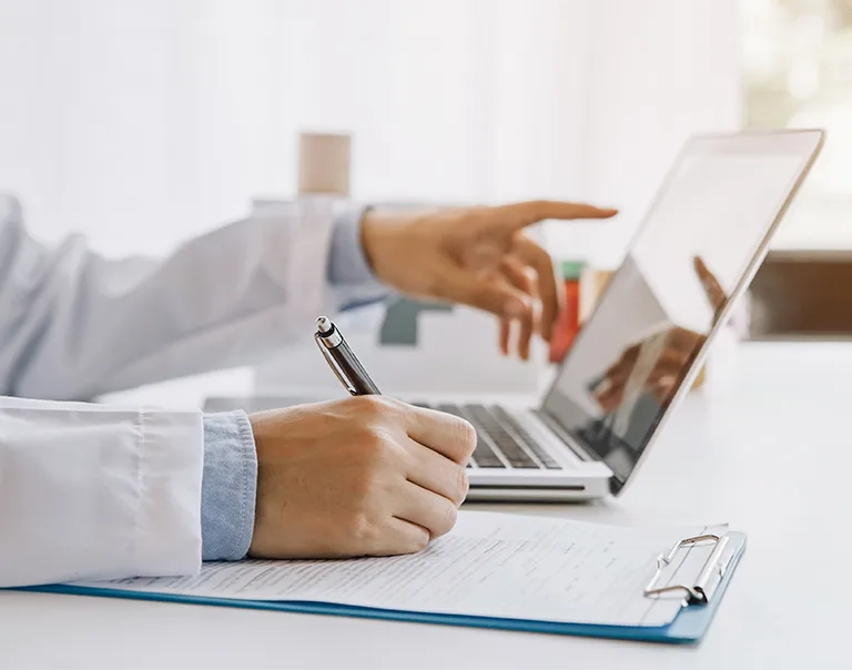 Image of a doctors hands writing on a notepad and pointing at a computer