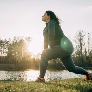 Overweight young woman warming up in morning sunlight before jogging outdoors. Weight losing, sports, healthy lifestyle