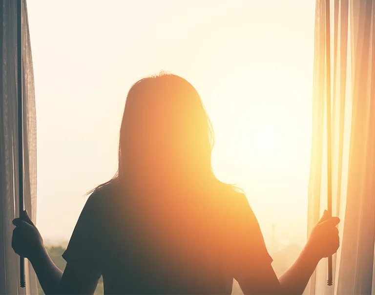 person standing in front of a window, opening the curtains to the morning sun
