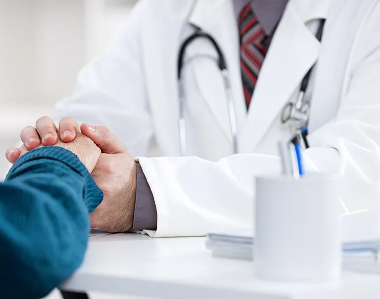 image of a doctor from the shoulders down, in a white coat with a stethoscope around their neck, reaching across a table and holding the hand of a patient with a blue shirt