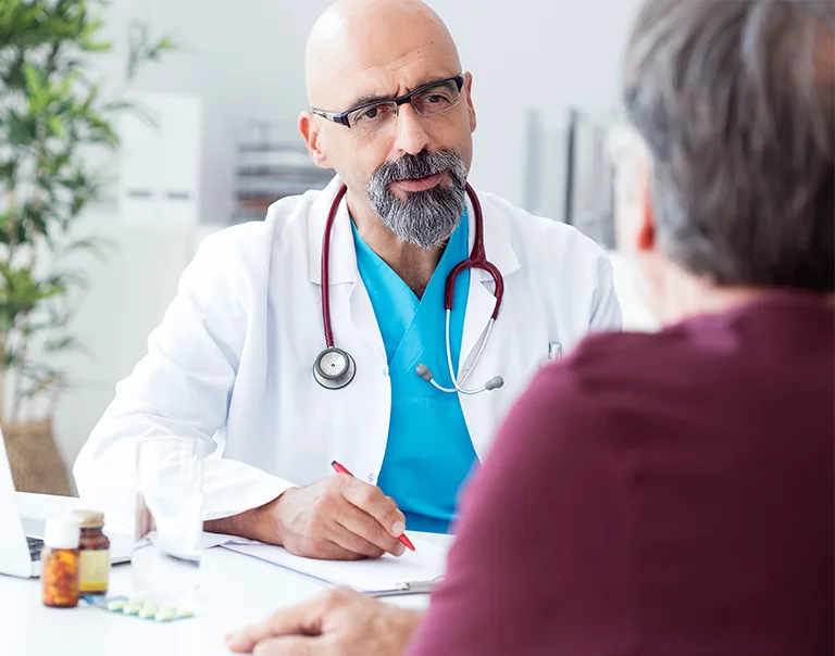 Image of a doctor taken from over the shoulder of a patient wearing a red shirt. The doctor is wearing a white coat with a blue shirt and has a stethoscope around their neck. They are speaking to the patient. The white OMA logo sits in the top right corner of the image.