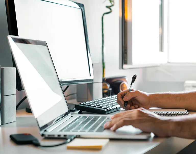 Image of a desk with a laptop and a monitor. There are hands typing on the laptop with a  pen and sticky notepad to the left of the laptop.
