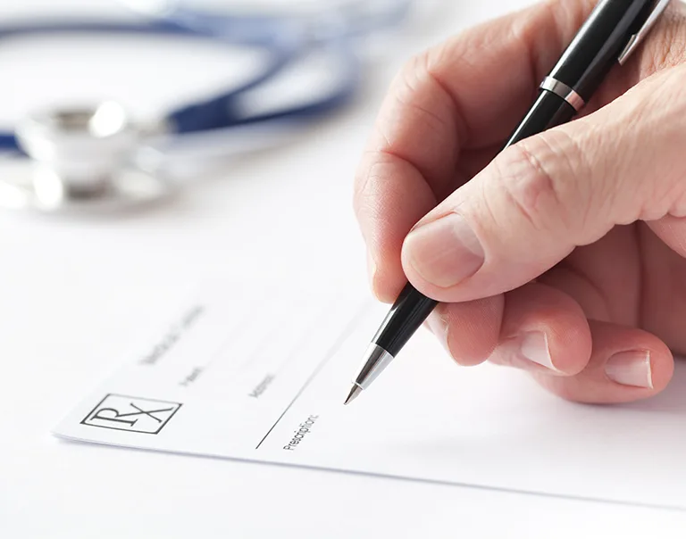 Close-up image of a hand with a pen hovering over a prescription pad with a stethoscope in the background
