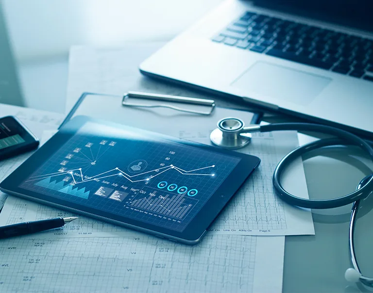 image of a desk with a computer, a stethoscope, an iPad with data and charts displayed and graph papers