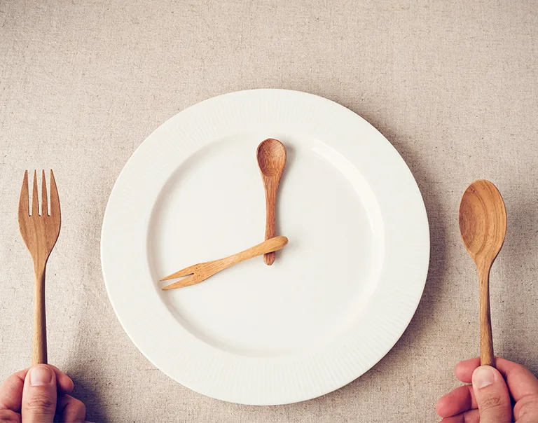 Image of a plate with a person holding a fork and spoon on either side of the plate. Another fork and spoon sit on the plate, to look like clock hands, making the plate look like a clock