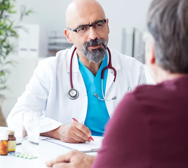Image of a doctor taken from over the shoulder of a patient wearing a red shirt. The doctor is wearing a white coat with a blue shirt and has a stethoscope around their neck. They are speaking to the patient. The white OMA logo sits in the top right corner of the image.