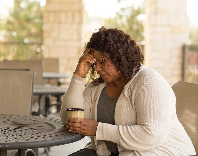 woman sitting on a patio at a table with a cup of coffee, with her head in her hands
