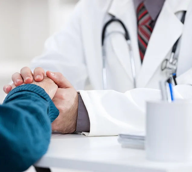 image of a doctor from the shoulders down, in a white coat with a stethoscope around their neck, reaching across a table and holding the hand of a patient with a blue shirt