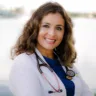 Headshot of Doris Munoz-Mantilla, MD, FOMA, DABOM, with a blurred lake in the background, wearing a white coat with a blue undershirt and a pink stethoscope around her neck.