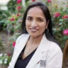 Headshot of Anila Chadha MD, DABOM, in a white coat posing in front of a rose bush