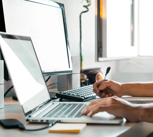 Image of a desk with a laptop and a monitor. There are hands typing on the laptop with a  pen and sticky notepad to the left of the laptop.