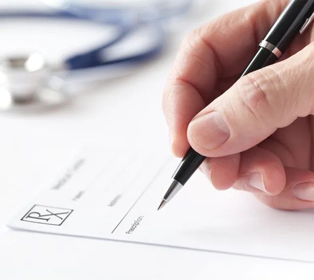 Close-up image of a hand with a pen hovering over a prescription pad with a stethoscope in the background
