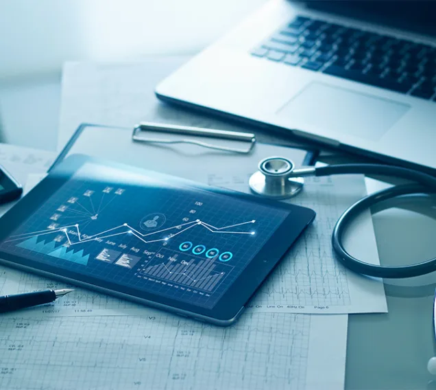 image of a desk with a computer, a stethoscope, an iPad with data and charts displayed and graph papers