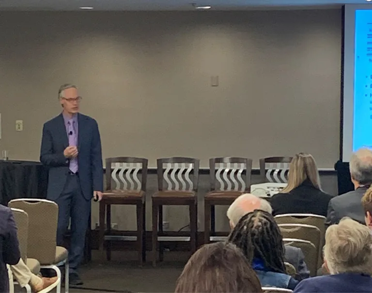 Image of Dr. Ethan Lazarus speaking in front of an audience in a conference room, with a presentation on the screen next to him